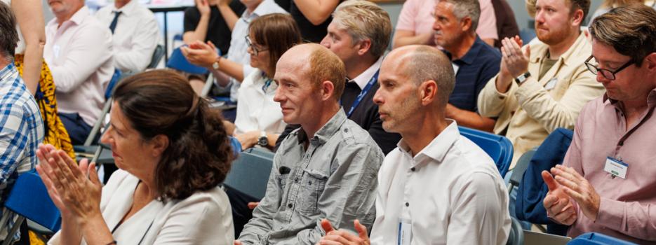 People of a range of ages in casual business clothes look ahead and clap at a green energy and innovation forum